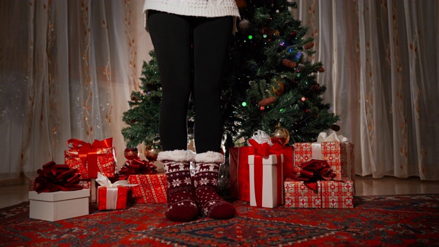 特写的苗条女人的腿在针织温暖的冬天袜子呆在旁边的圣诞树装饰。女孩的腿和圣诞树下的红色圣诞礼盒。树上闪烁的花环照亮了节日装饰的房间。家里的圣诞心情视频素材