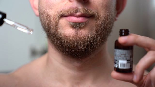 英俊的胡子男子呆在浴室的镜子前，拿着一个打开的玻璃瓶，里面有新鲜的芳香精油，用于软化干燥的男人剃须和刺激后的胡子。理发店的胡子护理产品，放松精油视频素材