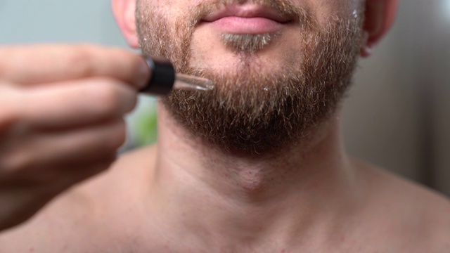 这是一个帅气的大胡子男子呆在浴室的镜子前，把新鲜的香精油滴在他干燥的胡子上的特写镜头。电动剃须器剃须后的放松程序。用于面部毛发治疗的精油视频素材