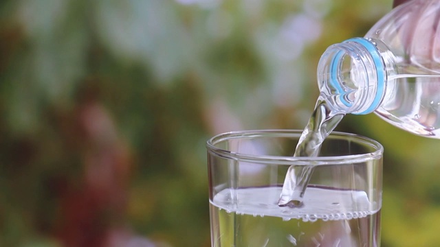 女人手握水瓶，将清澈的饮用水倒进模糊的绿色自然背景的玻璃杯中。视频素材