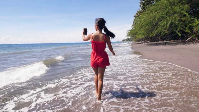 旅行女视频博主在海滩上散步，用智能手机录制视频。视频素材