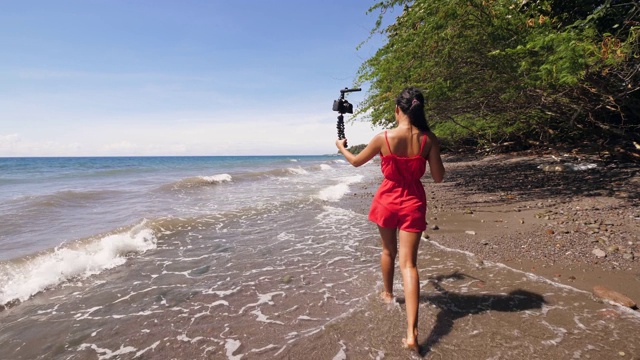 一个女视频博主在海滩上行走并录制自拍视频的后视图。视频素材