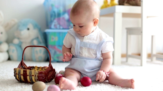 可爱的蹒跚学步的小男孩，在阳光明媚的客厅里玩着复活节巧克力兔子和五颜六色的复活节彩蛋视频素材