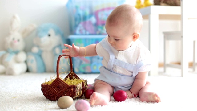 可爱的蹒跚学步的小男孩，在阳光明媚的客厅里玩着复活节巧克力兔子和五颜六色的复活节彩蛋视频素材