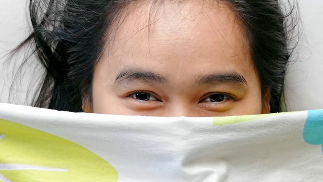 一名亚洲年轻女子早晨用毯子盖住自己的脸，脸上挂着微笑。视频素材