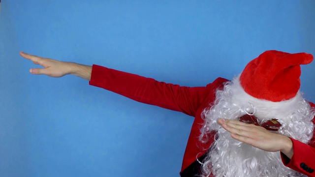 一个留着胡子，穿着圣诞老人服装的年轻人在蓝色背景上展示了现代姿态dab，节日视频下载