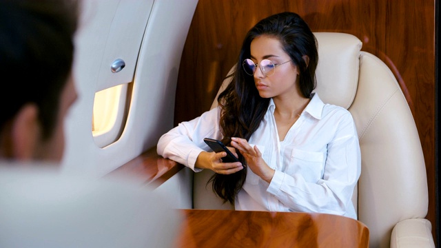 戴着眼镜、手持智能手机的迷人商务女性乘坐私人飞机旅行。商务旅行。穿着经典。女百万富翁乘坐商务舱的喷气式飞机。视频下载