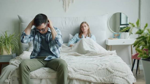 不开心的男人坐在床上触摸悲伤的脸，而愤怒的妻子在家里大喊大叫视频素材