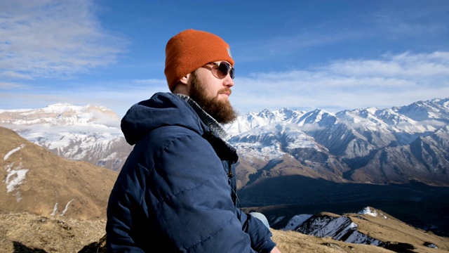 一个戴着太阳镜和帽子的大胡子旅行者的肖像坐在岩石上，背景是群山视频下载