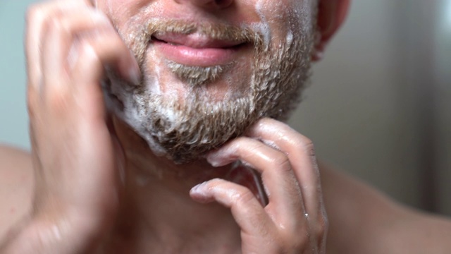 特写的年轻英俊的男子肥皂他的未剃胡须与特殊剃须洗发水从理发店洗它。早上例行野蛮男人在浴室，皮肤护理和胡须卫生，特别男性面部毛发治疗在家视频素材