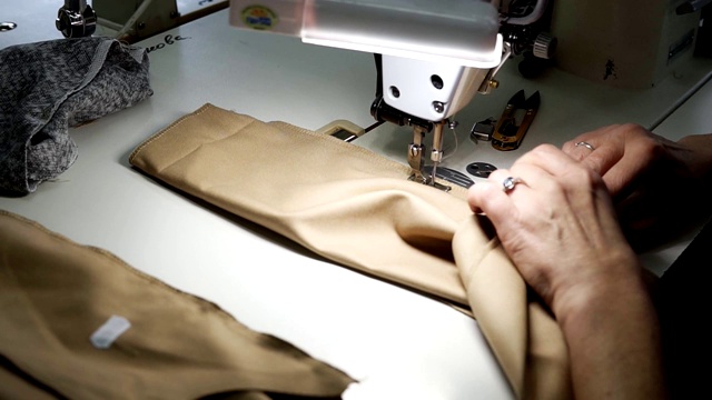 中年女性的双手在缝纫机上压脚和针附近工作。白人妇女时装设计师缝制米色面料样品在裁缝工作室。时尚和裁剪视频素材
