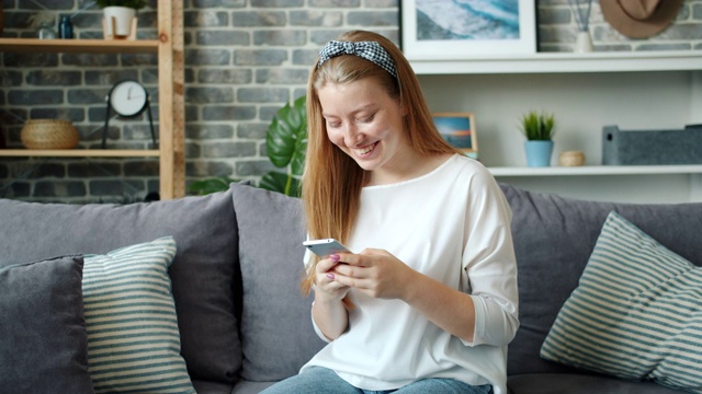 漂亮的金发女孩用着智能手机，在家里的沙发上放松地笑着视频素材