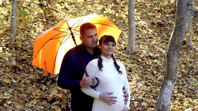 一对幸福的夫妇，一个怀孕的女孩和一个男人，站在一把橙色的雨伞下拥抱在一个晴朗的秋日。他们笑着，亲吻着，轻轻地抚摸着女孩的肚子。时尚的肖像。4 k。视频下载