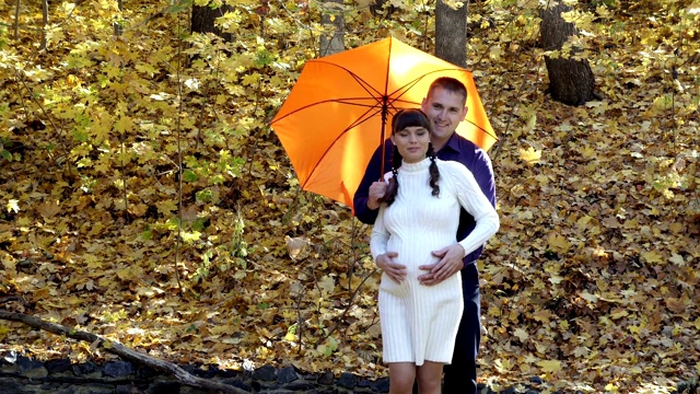 秋天的公园里，一个可爱的怀孕女孩和她的丈夫站在橙色的伞下。他们拥抱、玩耍、欢笑。一个穿白色针织连衣裙的女孩，一个穿蓝色衬衫的男人。特写镜头,4 k。视频下载