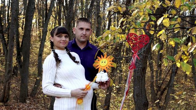 一对漂亮的夫妇，一个怀孕的女孩和一个男孩，在秋天的森林里，手里拿着白色的婴儿袜。在一个阳光明媚的日子里，他们在一片黄叶的背景下拥抱微笑。视频下载
