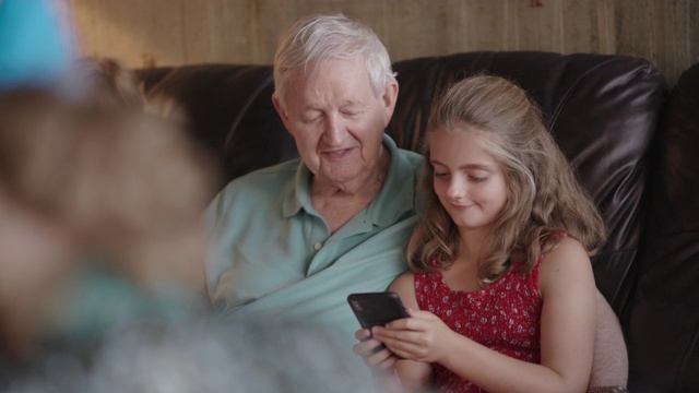 爷爷和孙女一起坐在客厅的沙发上，一边看着孙女的手机一边笑视频下载