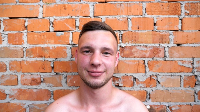 肖像微笑的年轻运动员裸体站在红砖墙的背景。这是一个长着大胡子的男人在室外直视镜头，脸上带着积极的情绪。视频下载