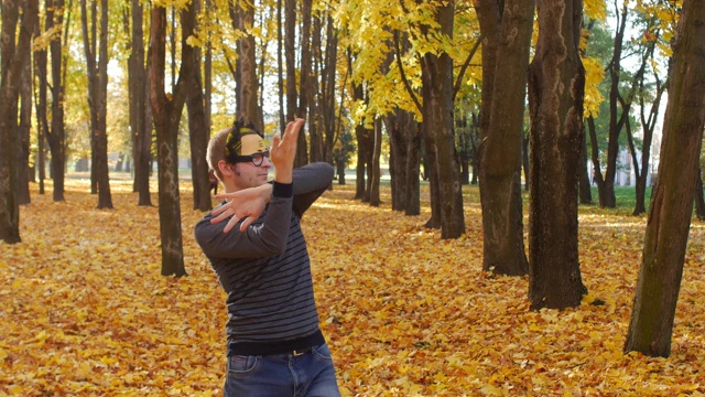 卷毛滑稽而快乐的人在秋天的公园里跳舞，滑稽而欢快的人类情感。一片树叶在阳光明媚的日子里飘落在一个小镇上。美丽的大自然视频素材