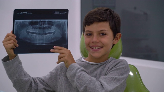 一个拉丁美洲男孩坐在牙医的椅子上，拿着一张牙齿的x光片对着镜头微笑视频素材