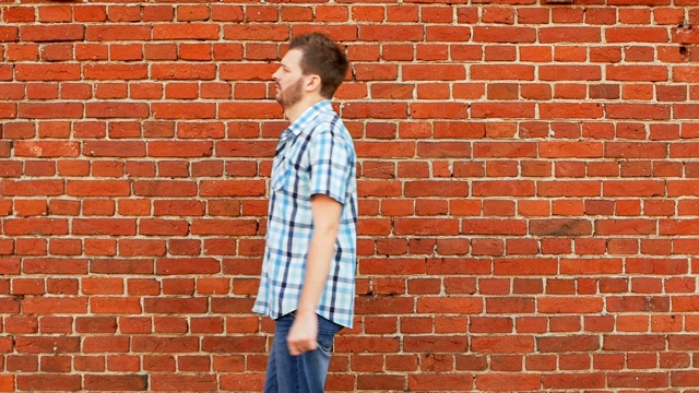 穿着衬衫和牛仔裤的有型胡须的男人沿着砖墙走视频素材