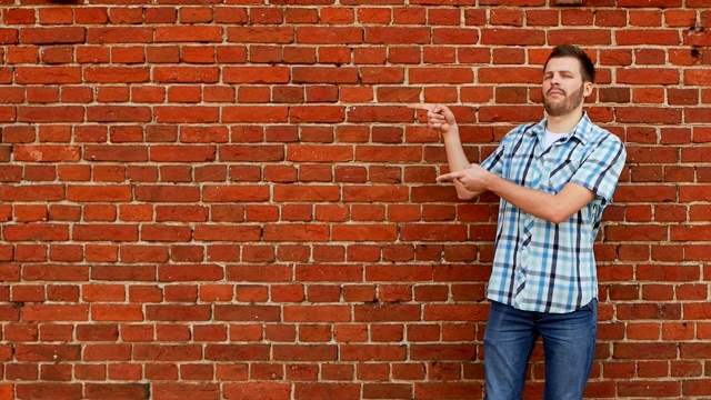 有型胡须的家伙穿着衬衫和牛仔裤指向复制空间，砖墙背景视频素材