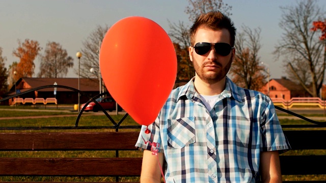 一个蓄着大胡子的家伙坐在长凳上，手里拿着一个红色的气球视频素材
