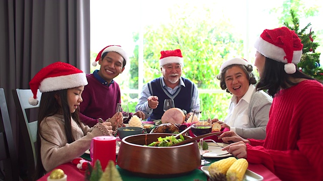 形形色色的亚洲人聚集在一起庆祝圣诞节。头发花白的爷爷奶奶，兄弟姐妹，儿子，女儿参加家庭宴会，坐在自制的午餐桌旁，在冷杉树下交谈。视频素材