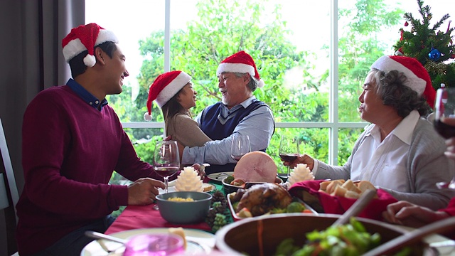 形形色色的亚洲人聚集在一起庆祝圣诞节。头发花白的爷爷奶奶，兄弟姐妹，儿子，女儿参加家庭宴会，坐在自制的午餐桌旁，在冷杉树下交谈。视频素材
