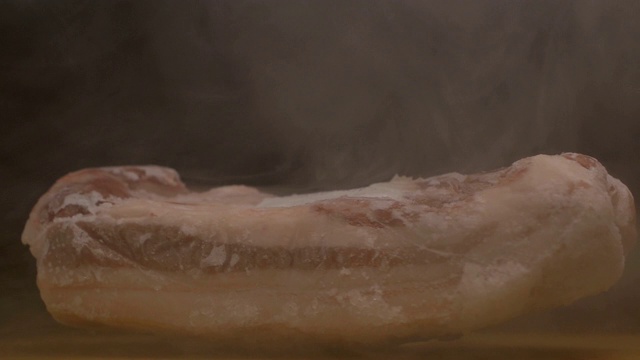 一块冷冻旋转的猪肉在黑色的背景下，霜冻的新鲜感和烟雾从肉中传来，特写，慢墨视频下载