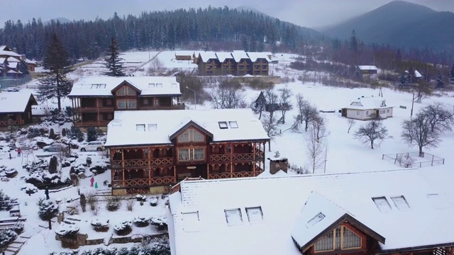 在被针叶林包围的山村木制小屋的空中。在滑雪胜地被雪覆盖的小屋的无人机视图。寒冷的冬日，山上下雪视频素材