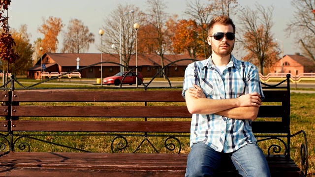 留着胡子、戴着太阳镜、穿着衬衫和牛仔裤的时髦男人坐在长凳上视频素材