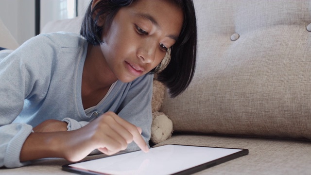 亚洲小女孩躺在家里客厅的沙发上，触摸着白色的数字平板电脑屏幕，微笑着。视频素材