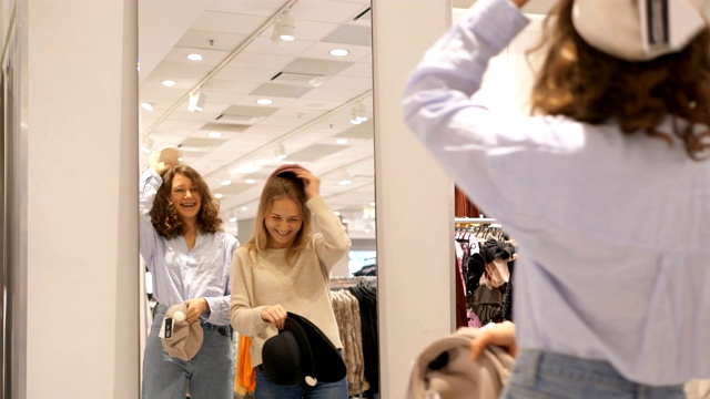 有趣的闺蜜在服装店的镜子前试帽子，然后大笑视频下载
