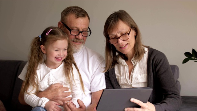 家庭、一代、技术和人的概念——微笑的祖父、孙女和祖母坐在家里的沙发上，拿着平板电脑视频素材