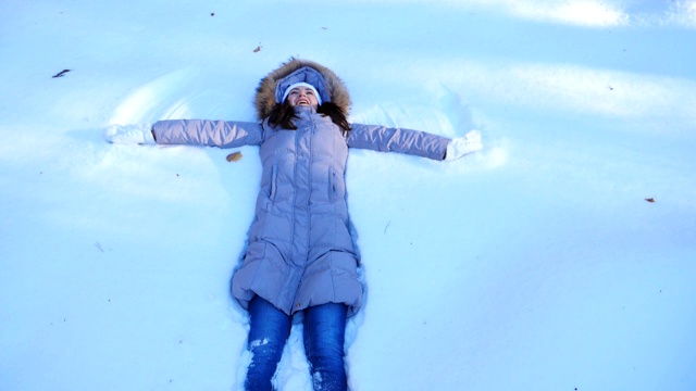 年轻的女孩躺在白雪覆盖的草地上做天使。快乐的女人在雪地里玩，享受冬天的时光。快乐的女士在户外玩。寒假的概念。慢镜头视频素材