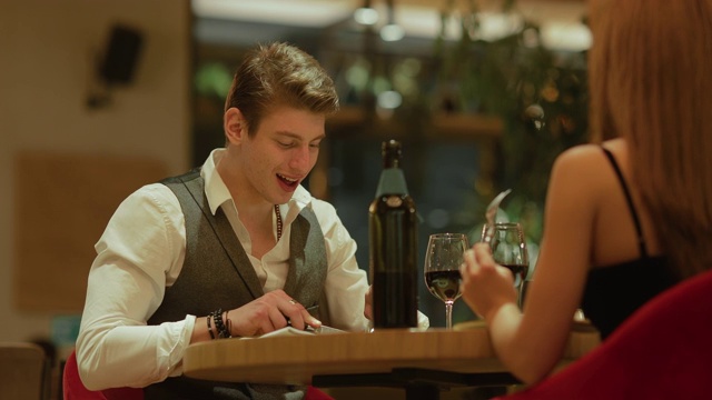 一个年轻人在餐桌上和一个女孩聊天。英俊的男人吃喝，在一个舒适的餐厅约会。视频下载