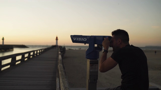 8月30日，法国多维尔，一名男子在海滩上用望远镜欣赏美丽的橙色日落。黑色衬衫，棕色头发和胡子。4 k UHD。视频下载