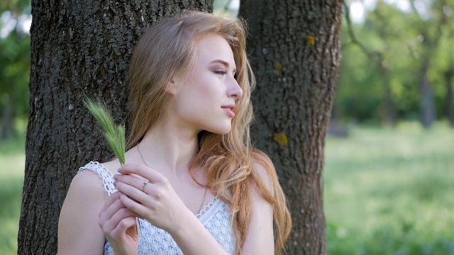 一个漂亮的金发女孩的肖像。缓慢的运动。那个女孩正在公园里休息。在公园里，一个女孩坐在树旁的草地上视频素材