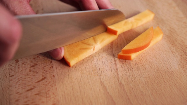 一个年轻人的手把一把刀切成小块，一个成熟的橘子柿子放在一块木头砧板上视频下载