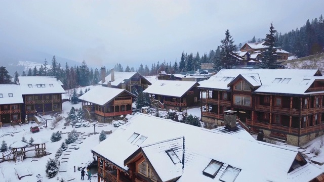 在被针叶林包围的山村木制小屋的空中。在滑雪胜地被雪覆盖的小屋的无人机视图。寒冷的冬日，山上下雪视频下载