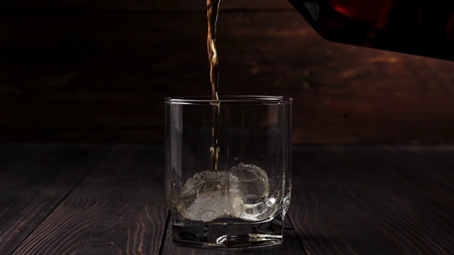 奢侈威士忌。Hand从一个方形瓶子里倒出金色的威士忌，倒进一个有真正冰的玻璃杯，放在桌子上，背景是木制的。苏格兰在滚筒。在高杯酒波旁威士忌。视频素材