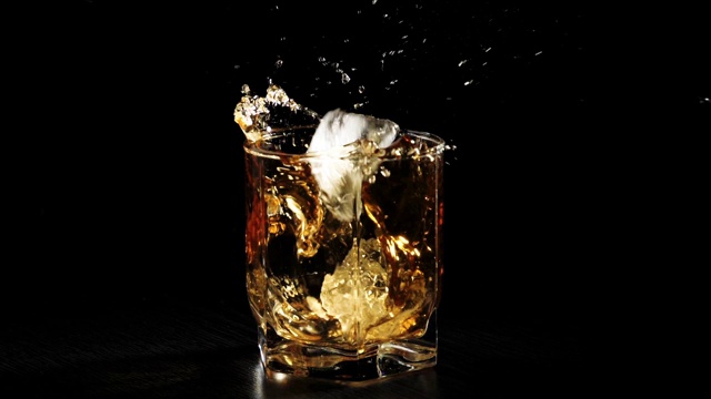 奢侈威士忌。在黑色背景下的黑色桌子上，冰块掉落在威士忌杯中，溅起冰块。苏格兰在滚筒。在高杯酒波旁威士忌。缓慢的运动。视频素材
