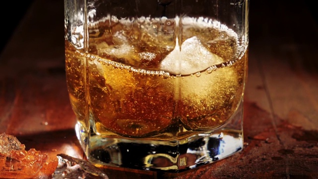 奢侈威士忌。冰块飞溅到棕色木桌上的一只盛着金色威士忌的玻璃杯里。苏格兰在滚筒。在高杯酒波旁威士忌。缓慢的运动。视频素材