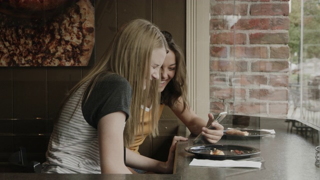 美国犹他州普罗沃，女孩们坐在披萨店的窗口边用手机查看社交媒体视频素材