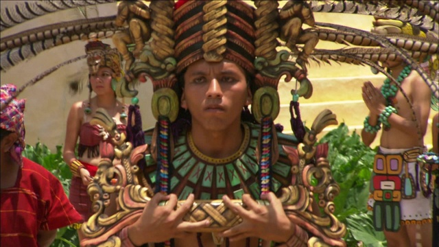 一个戴着羽毛头饰、穿着玛雅服装的男人打着蝴蝶结，走在旁观者身边。视频下载
