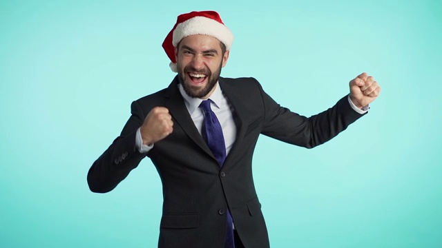 肖像英俊的商人在西装和圣诞老人帽跳舞孤立在蓝色演播室背景。年轻快乐的男人胡子庆祝圣诞或新年派对。视频素材