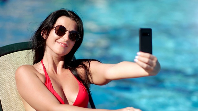 微笑的时尚女性戴着时髦的太阳镜，在海水背景下用智能手机自拍视频素材