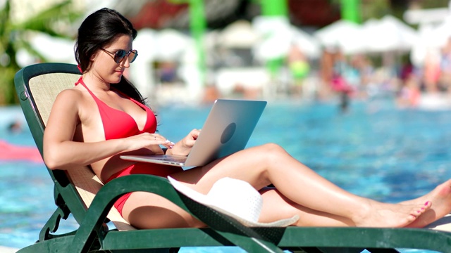穿着红色泳衣的自由职业者女孩躺在游泳池附近的躺椅上用笔记本电脑工作视频素材