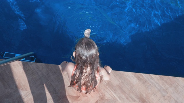 女孩坐在泳池边摇摆双腿，在泳池外俯视图。顶视图女孩溅腿在蓝色的水游泳池在度假酒店。视频素材