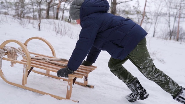 在下雪的冬天，穿着暖和衣服的男孩推着老式的木制雪橇跑上小山。一个在公园里滑雪的小家伙。森林里穿着海军夹克雪橇的孩子。慢动作视频素材
